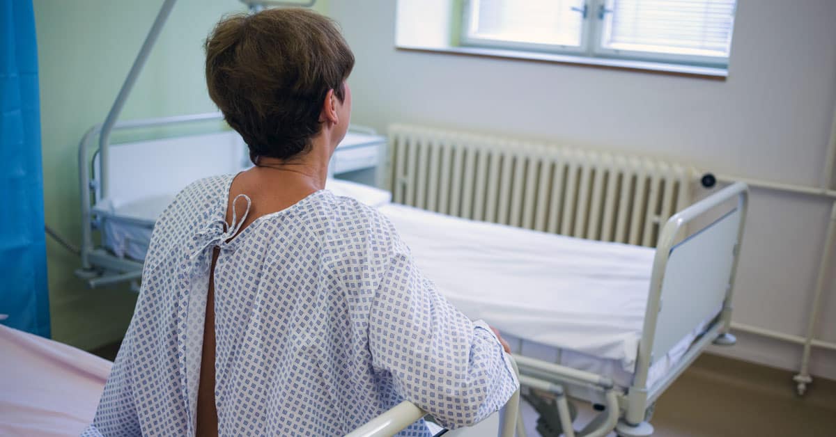 Lady sitting on edge of hospital bed with backside showing through hospital gown