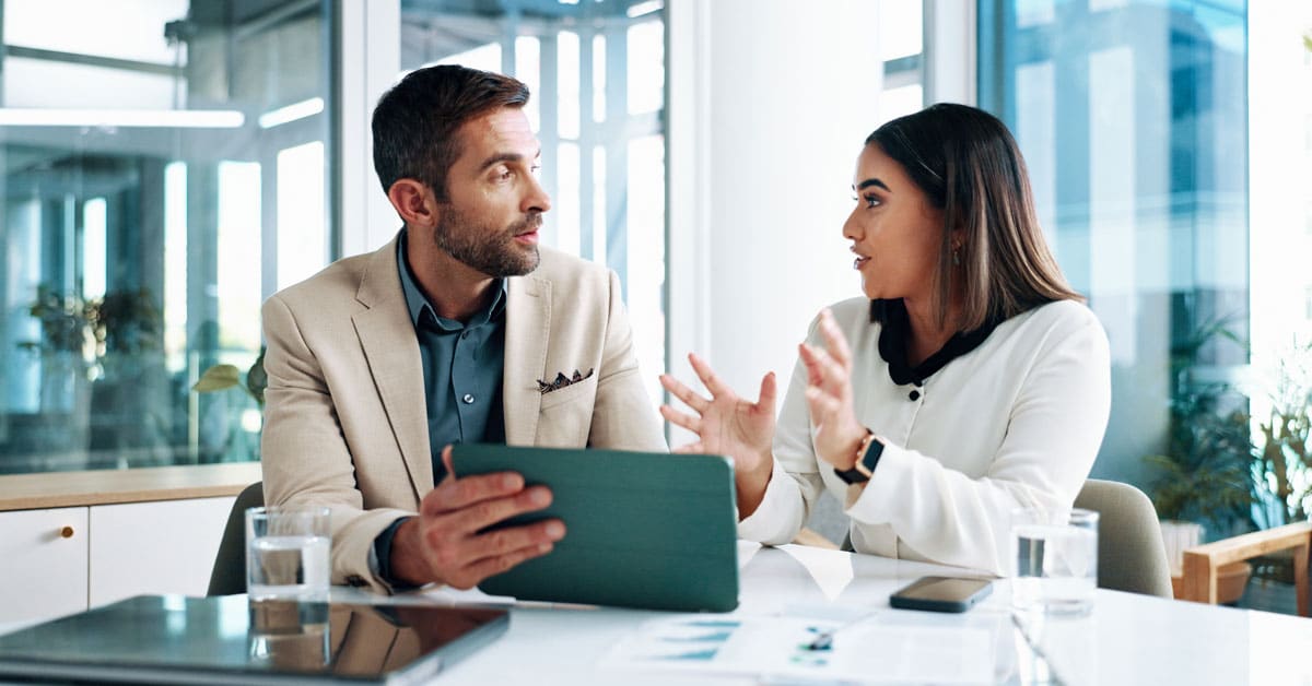 Two colleagues in a bright, modern office discuss at a clean table, with a tablet between them as they review a document together.