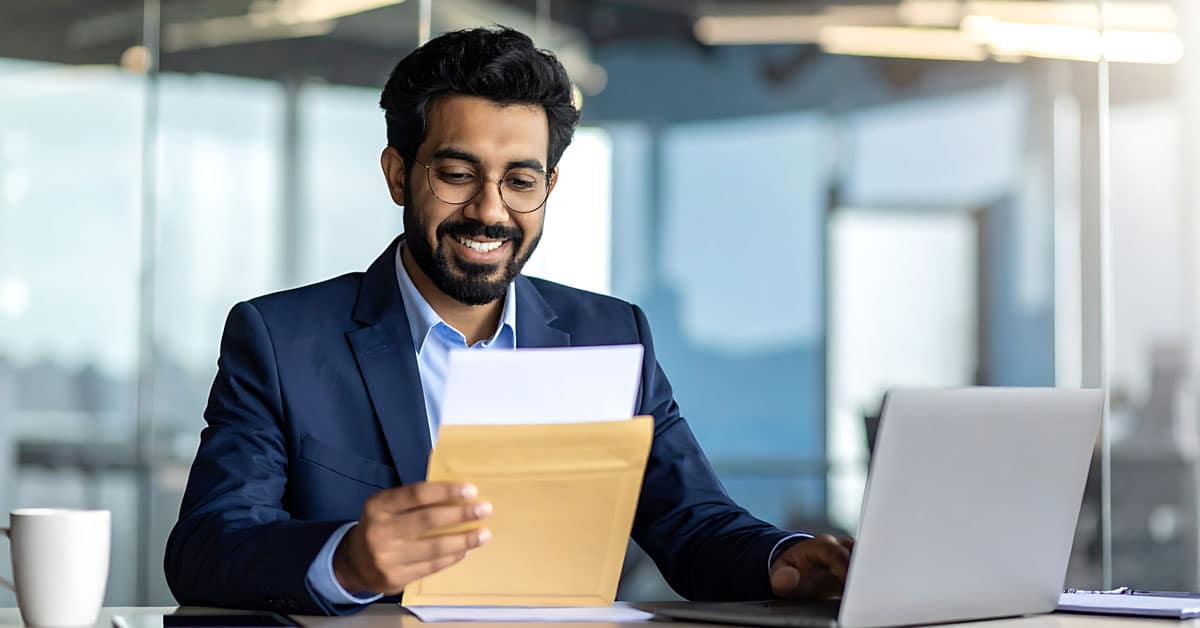 Smiling man in a navy suit reads a letter at his desk, with a laptop nearby in a modern office.